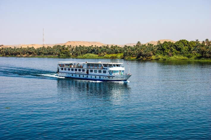 Cruise boat sailing on the Nile River past palm trees and desert landscape in Egypt