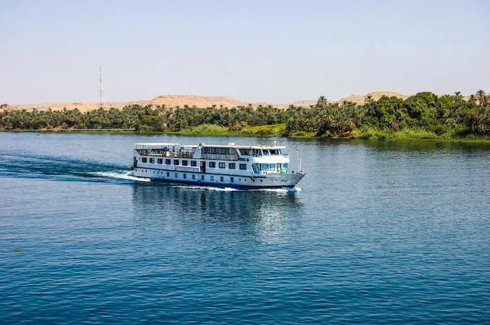 Cruise boat sailing on the Nile River past palm trees and desert landscape in Egypt