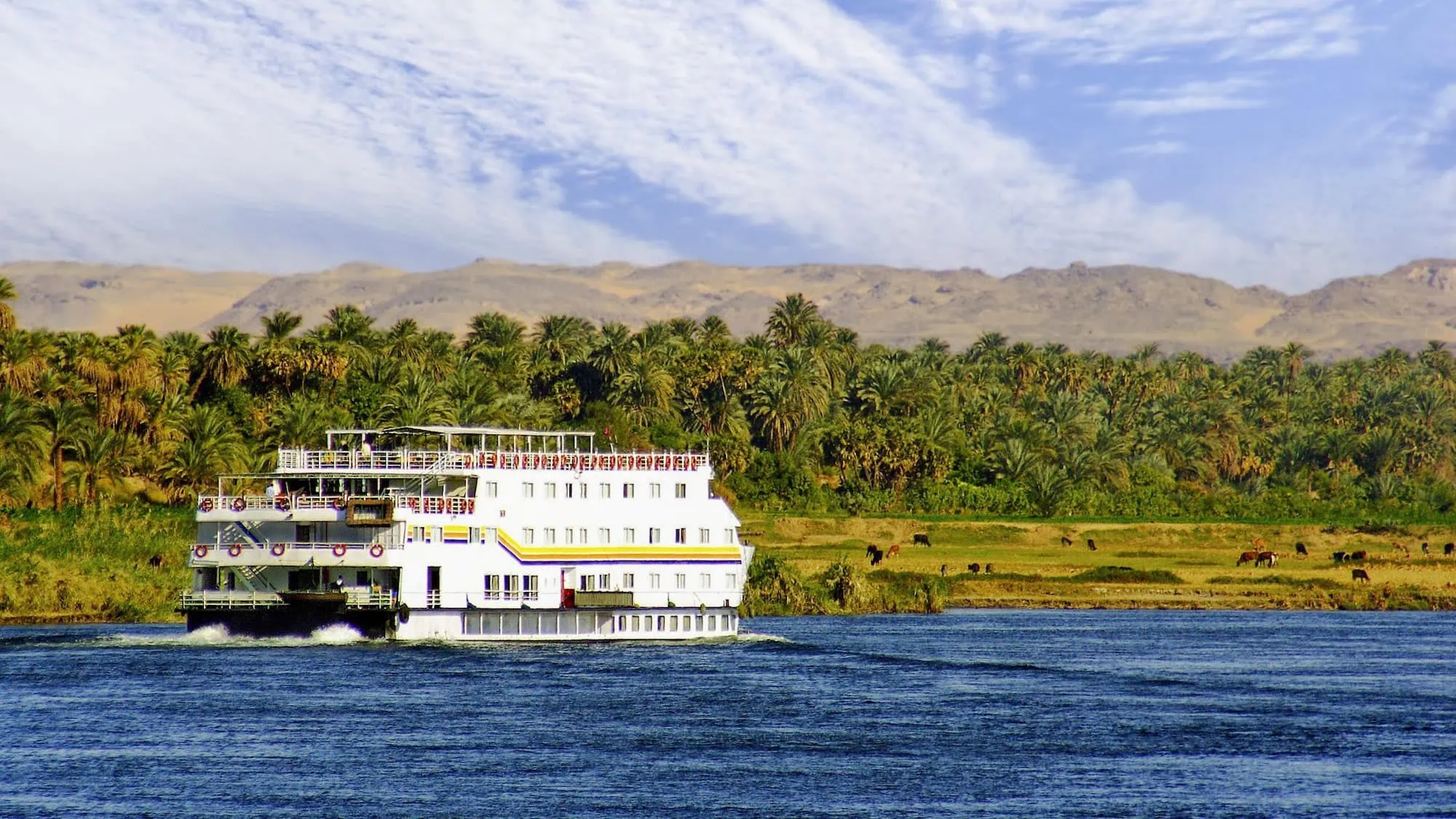White multi-deck cruise ship sailing on the Nile River with lush green riverbanks and desert mountains in background