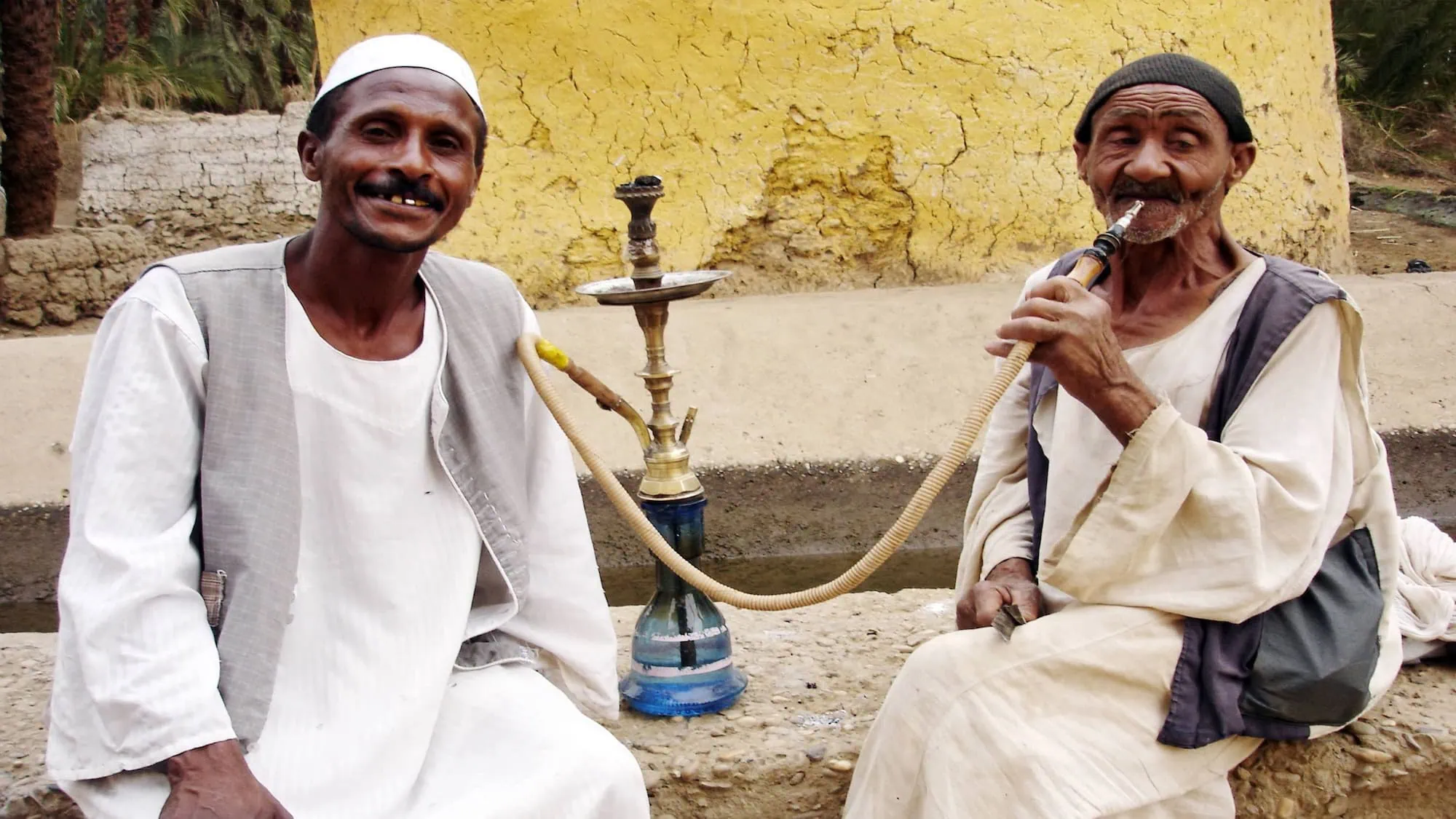 Two Nubian men in traditional white clothing smoking hookah in a village setting