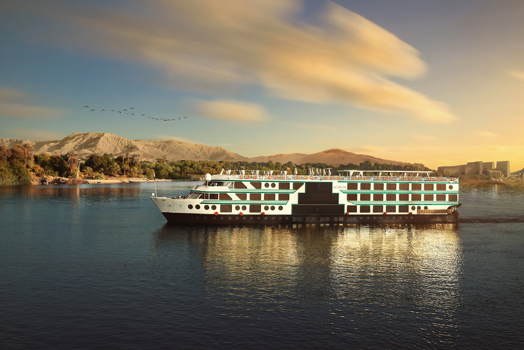 Nile River cruise ship sailing past desert mountains and palm trees under cloudy skies