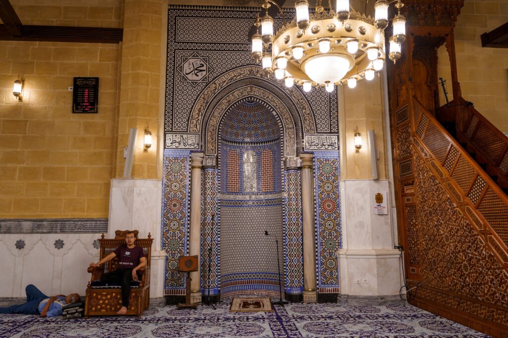 The mihrab area with decorative tilework, Arabic calligraphy, chandeliers, and carved wooden minbar inside Al-Hussein Mosque, Cairo