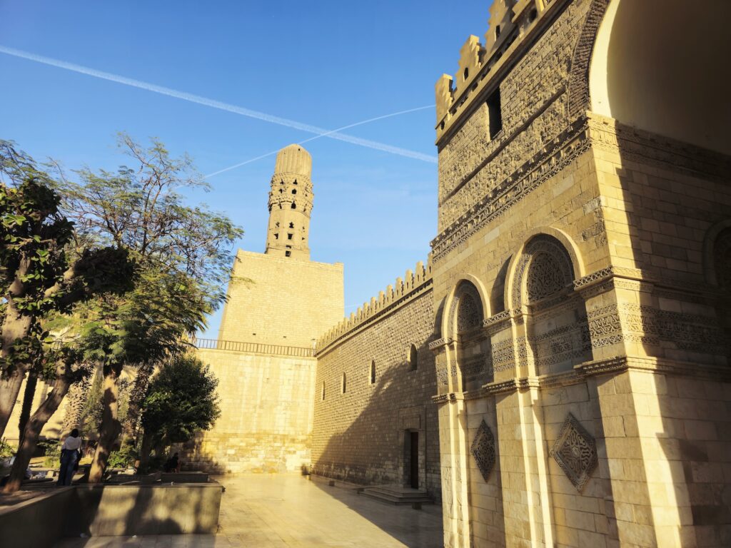 An exterior view showing the stone facade, entrance towers, and minarets of the Al-Hakim bi-Amr Allah Mosque along Al-Muizz Street near Bab al-Futuh, Cairo