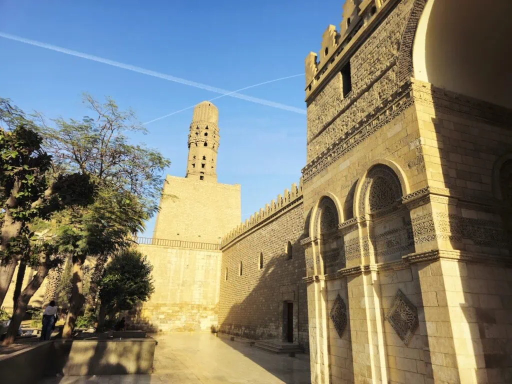 An exterior view showing the stone facade, entrance towers, and minarets of the Al-Hakim bi-Amr Allah Mosque along Al-Muizz Street near Bab al-Futuh, Cairo