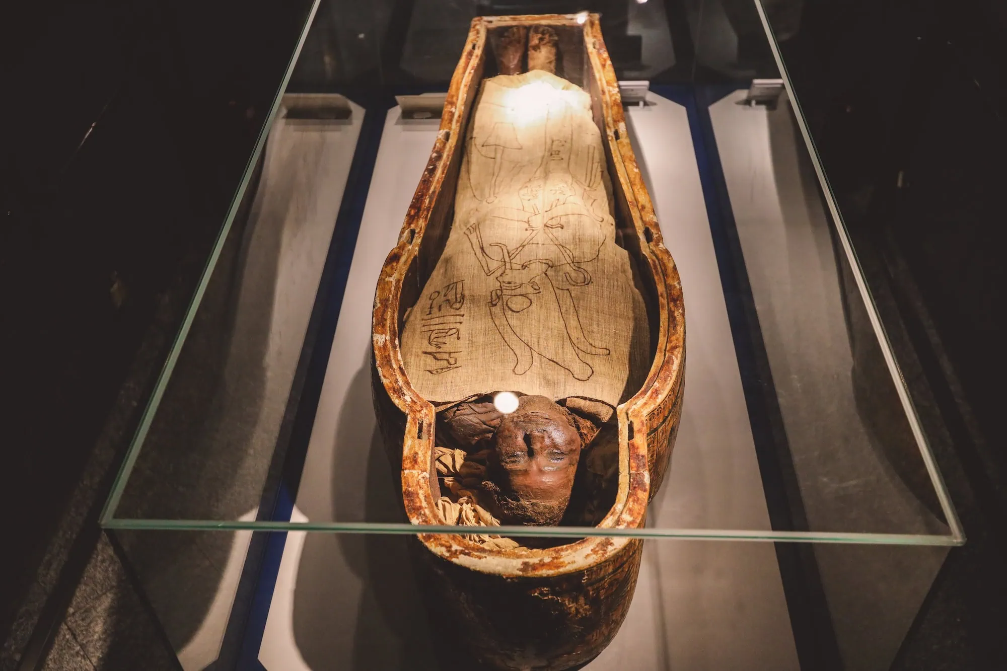 Ancient Egyptian sarcophagus with hieroglyphic decorations and preserved mummy displayed in museum case