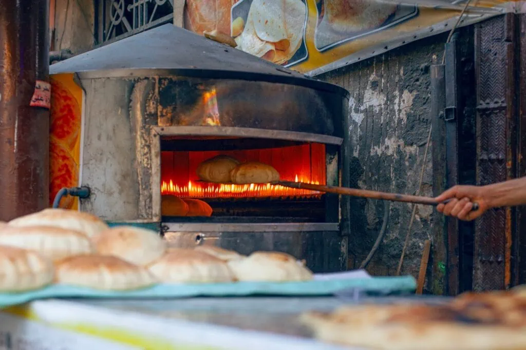 Egyptian baker placing rounds of aish baladi flatbread into a traditional oven inside a small street bakery