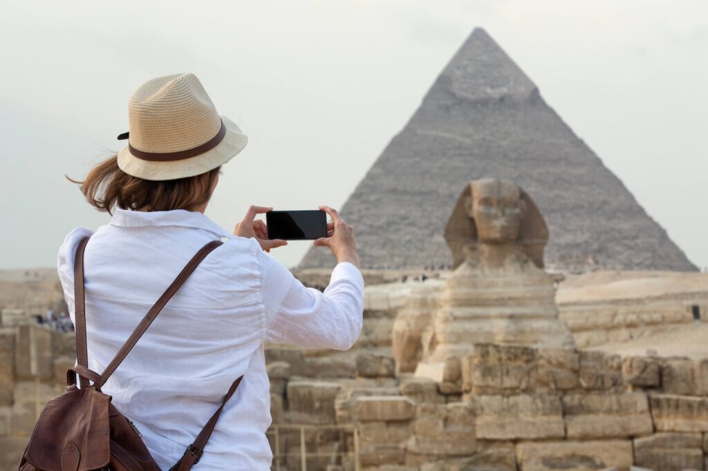Woman standing and taking a photo in front of the Great Sphinx with surrounding desert and pyramid structures visible, Giza