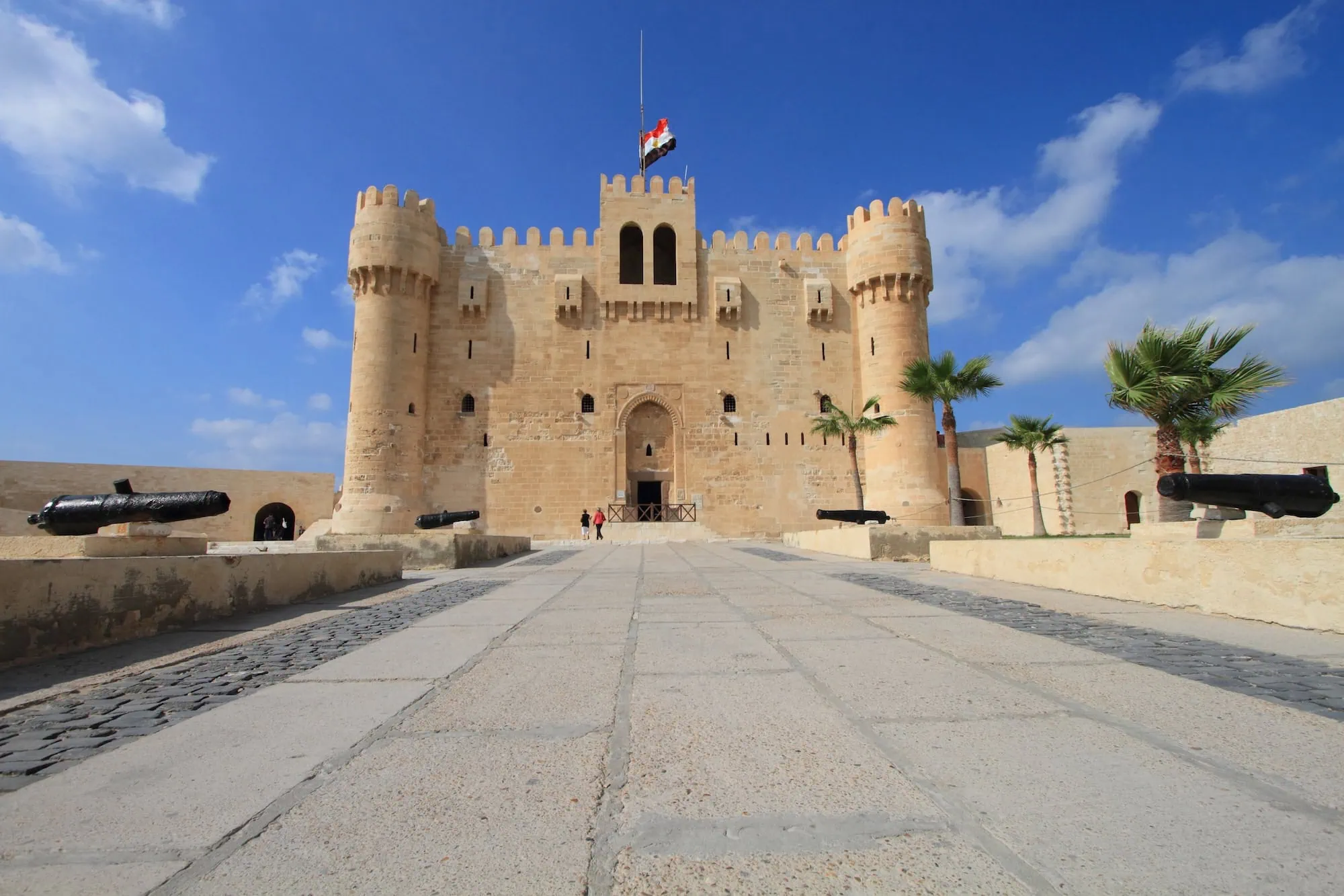 Qaitbay Citadel fortress in Alexandria with stone towers, Egyptian flag, palm trees and historic cannons