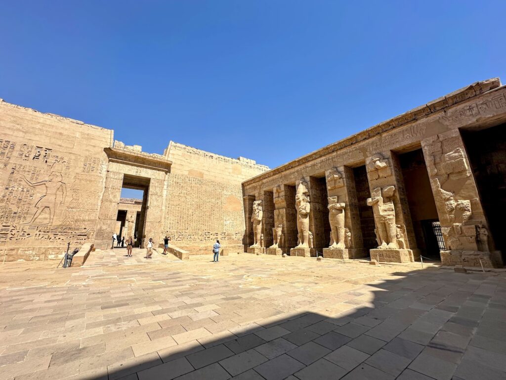Courtyard with Osiride statues, carved stone walls, and visitors inside the Medinet Habu temple complex, Luxor
