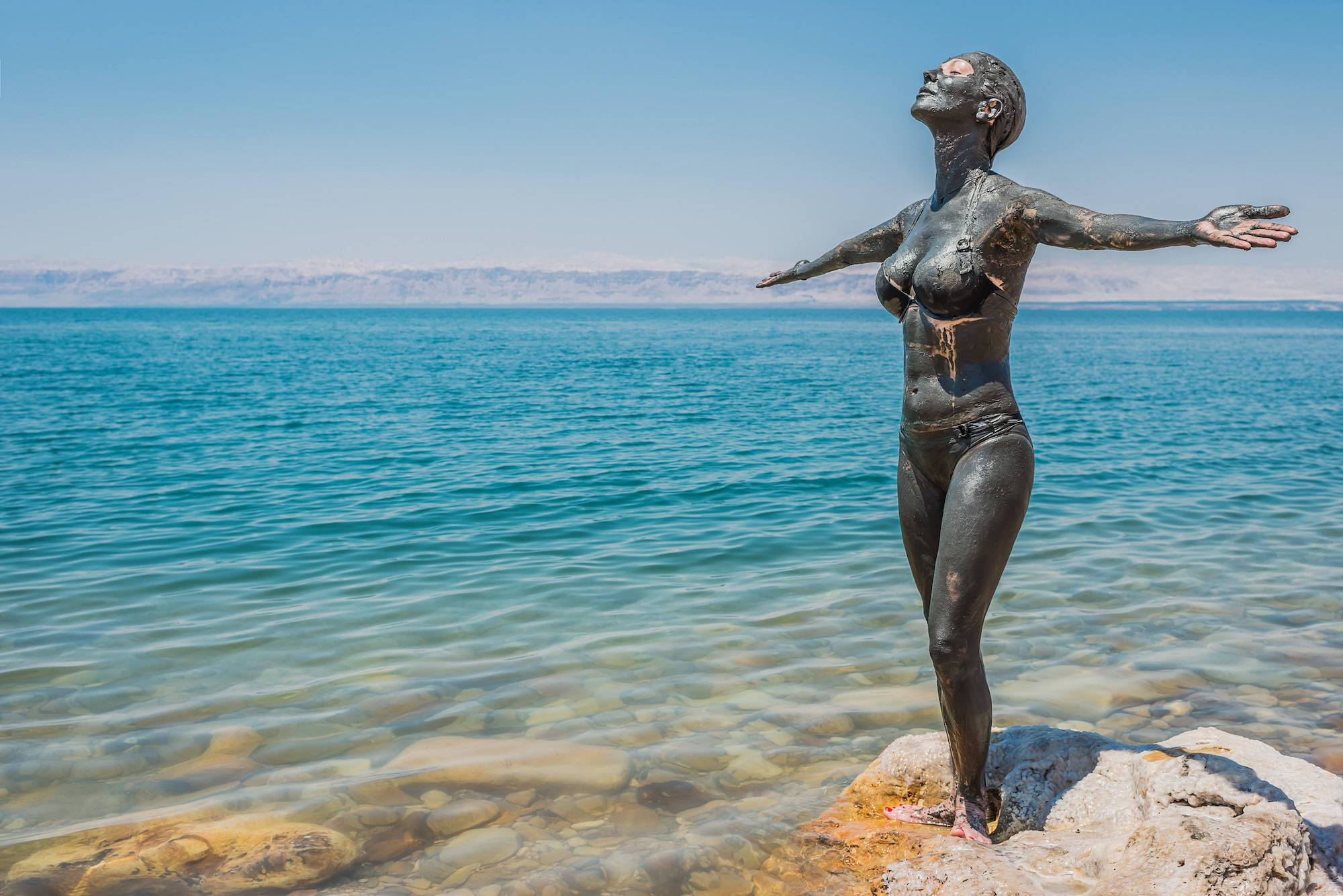 Person applying healing Dead Sea mud treatment with salt formations and mountains in background