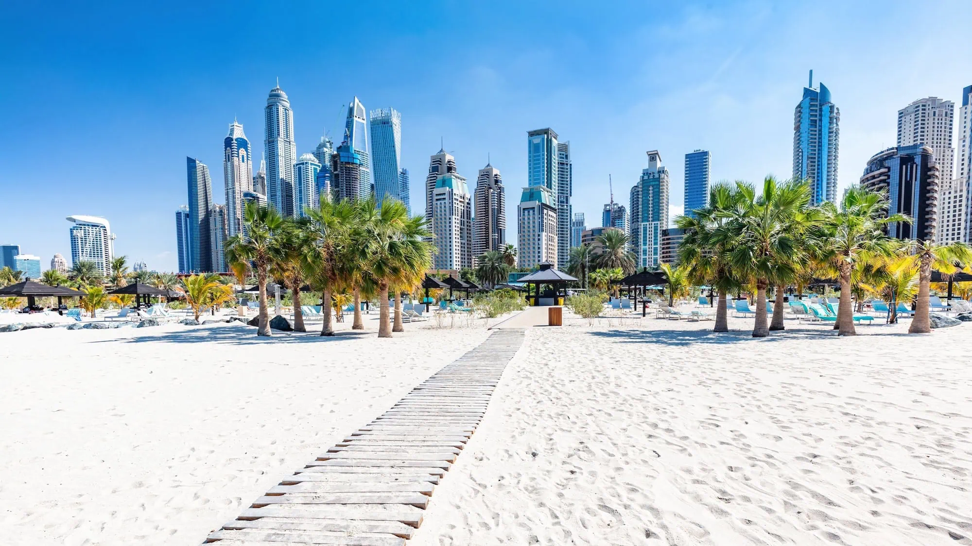 Jumeirah Beach with wooden boardwalk, palm trees, and Dubai Marina skyline in background