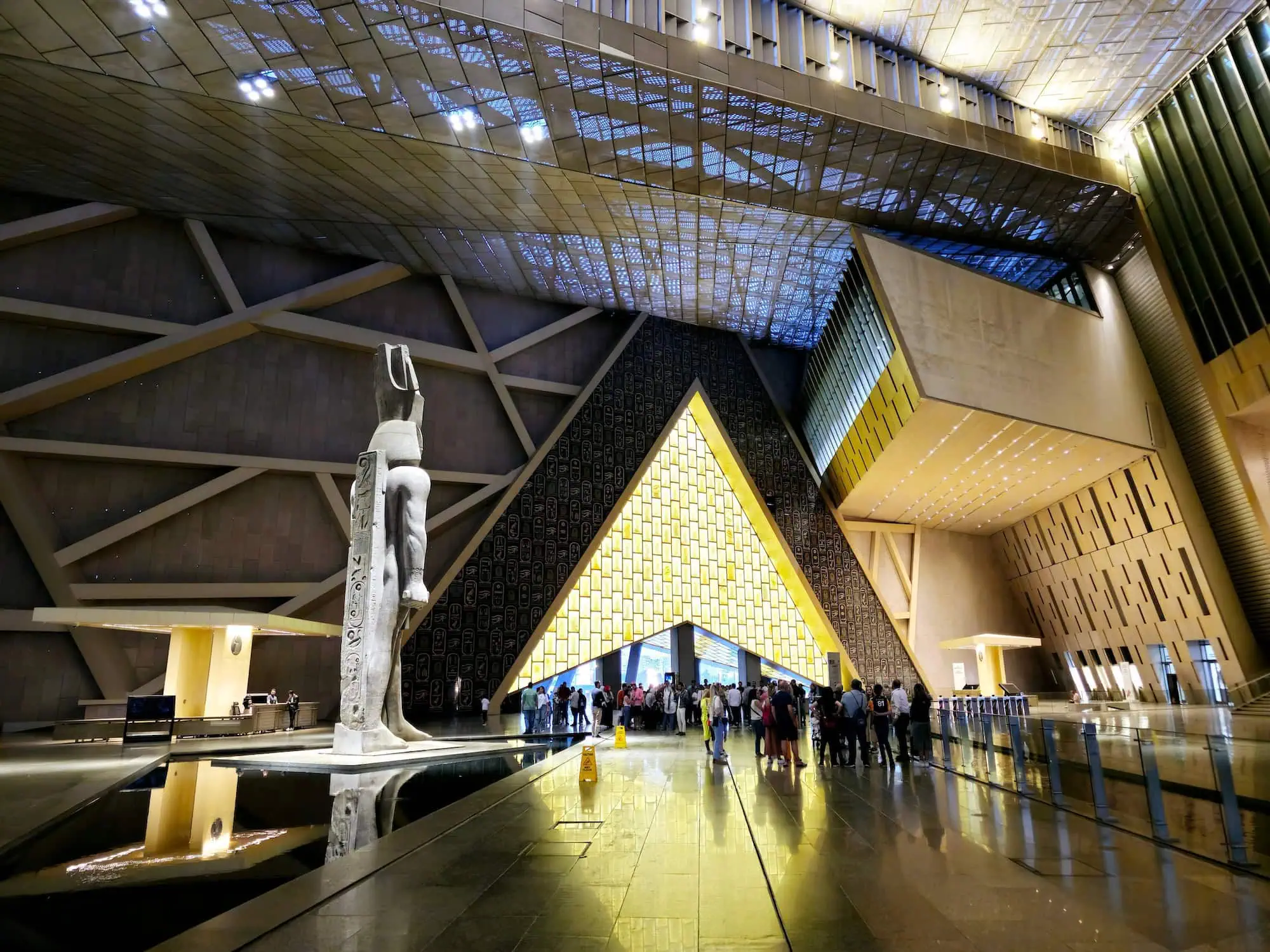 Interior view of Grand Egyptian Museum's main gallery with ancient statues, glass ceiling, and visitors