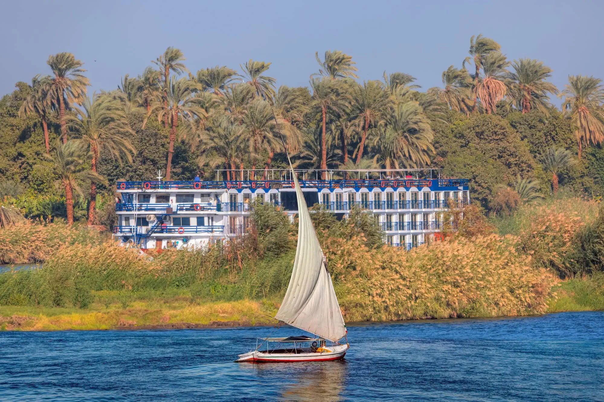 Felucca sailboat with white sail on Nile River during golden sunset with cruise ship in background