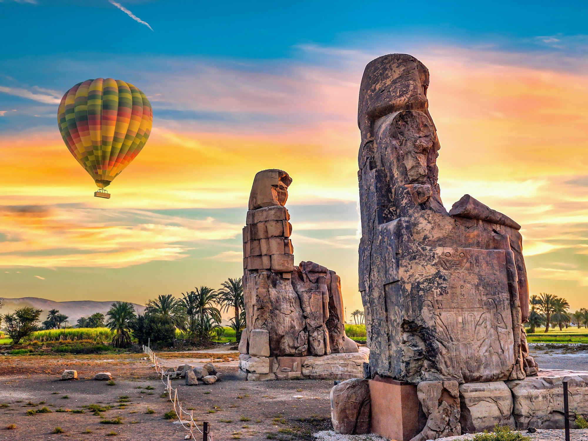 Hot air balloon floating above the ancient Colossi of Memnon statues in Luxor's desert landscape