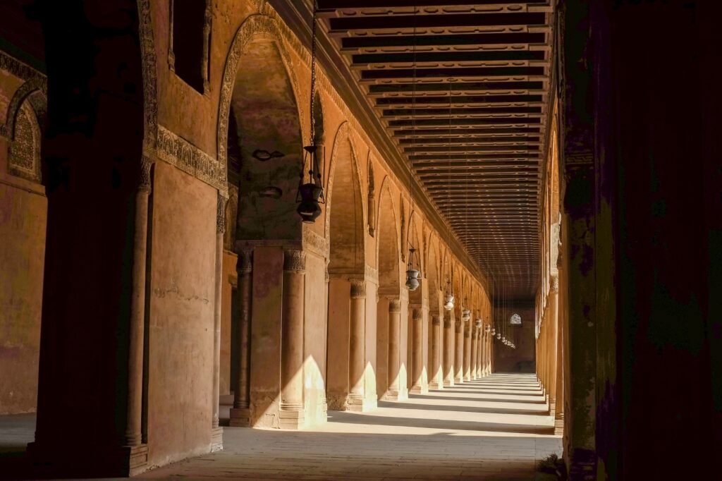 An arcaded corridor with pointed arches, stone columns, hanging lamps, and wooden ceiling at the Ibn Tulun Mosque, Cairo