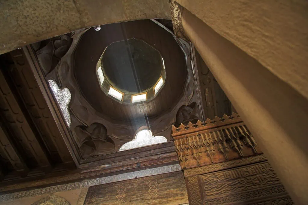 An interior view looking up at the central dome with wooden ceiling panels, carved details, and filtered daylight inside the Ibn Tulun Mosque, Cairo