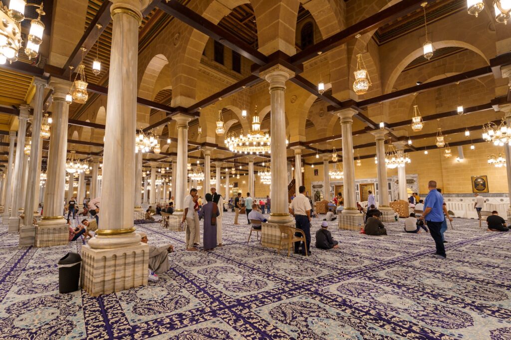 Worshippers praying among columns, arches, and hanging chandeliers inside Al-Hussein Mosque