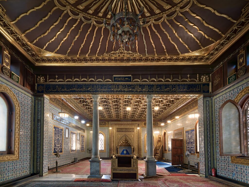 An ornate interior hall with carved wooden ceiling, patterned tile walls, marble columns, chandeliers, and Arabic calligraphy at Manial Palace, Cairo