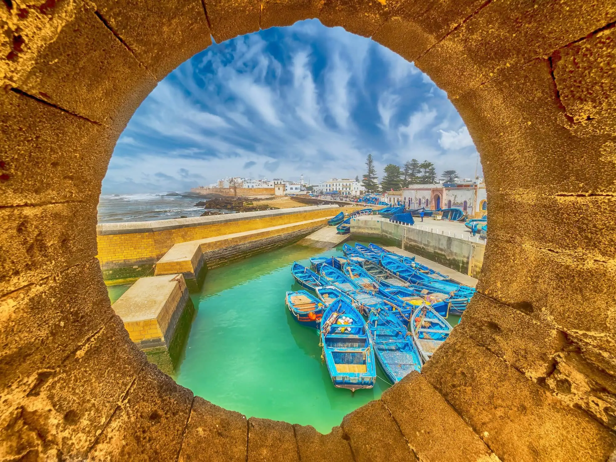 View of Essaouira's fishing harbor with blue boats and medina walls seen through fortress archway