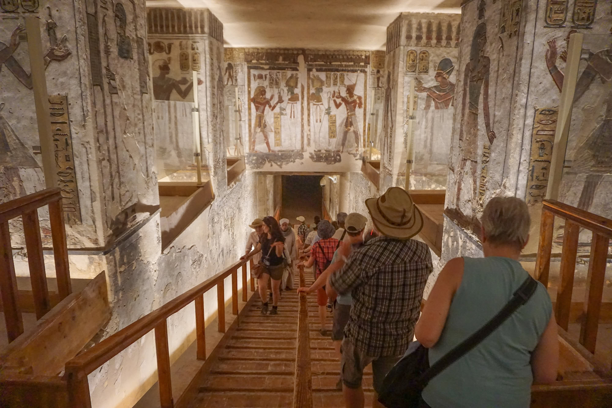 Tourists descending wooden stairs into ancient Egyptian tomb with hieroglyphic murals