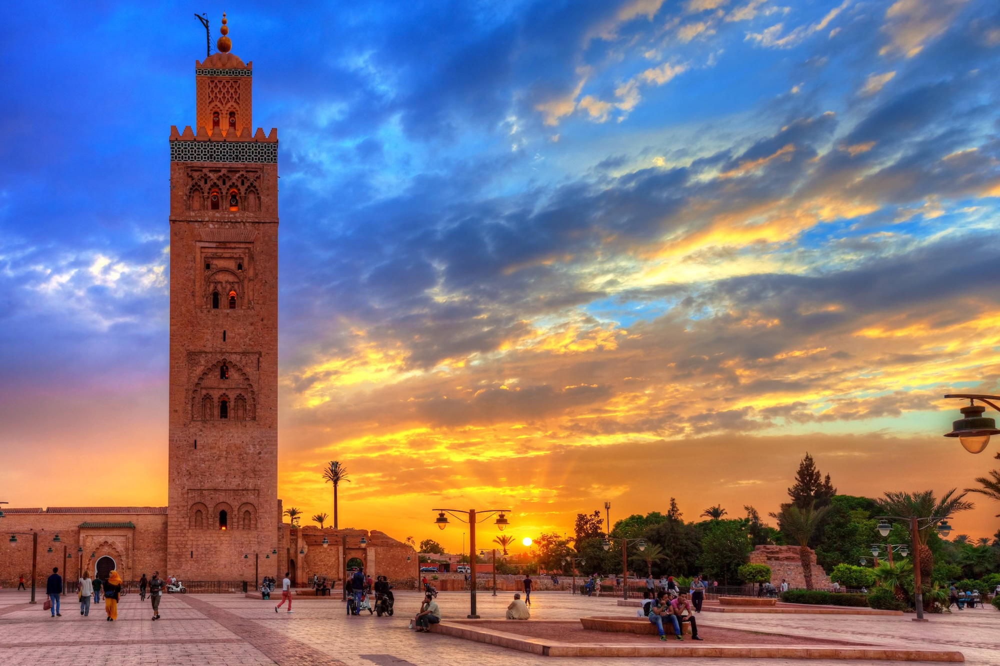 Tourists visiting Koutoubia Mosque in Marrakech with its towering minaret and palm trees at sunset