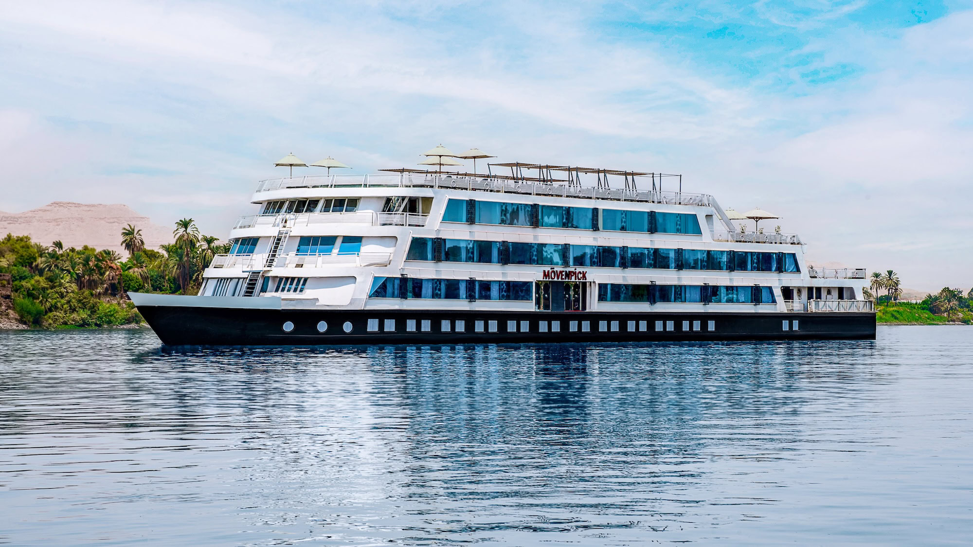 Nile River cruise ship sailing through serene waters with palm trees and desert hills