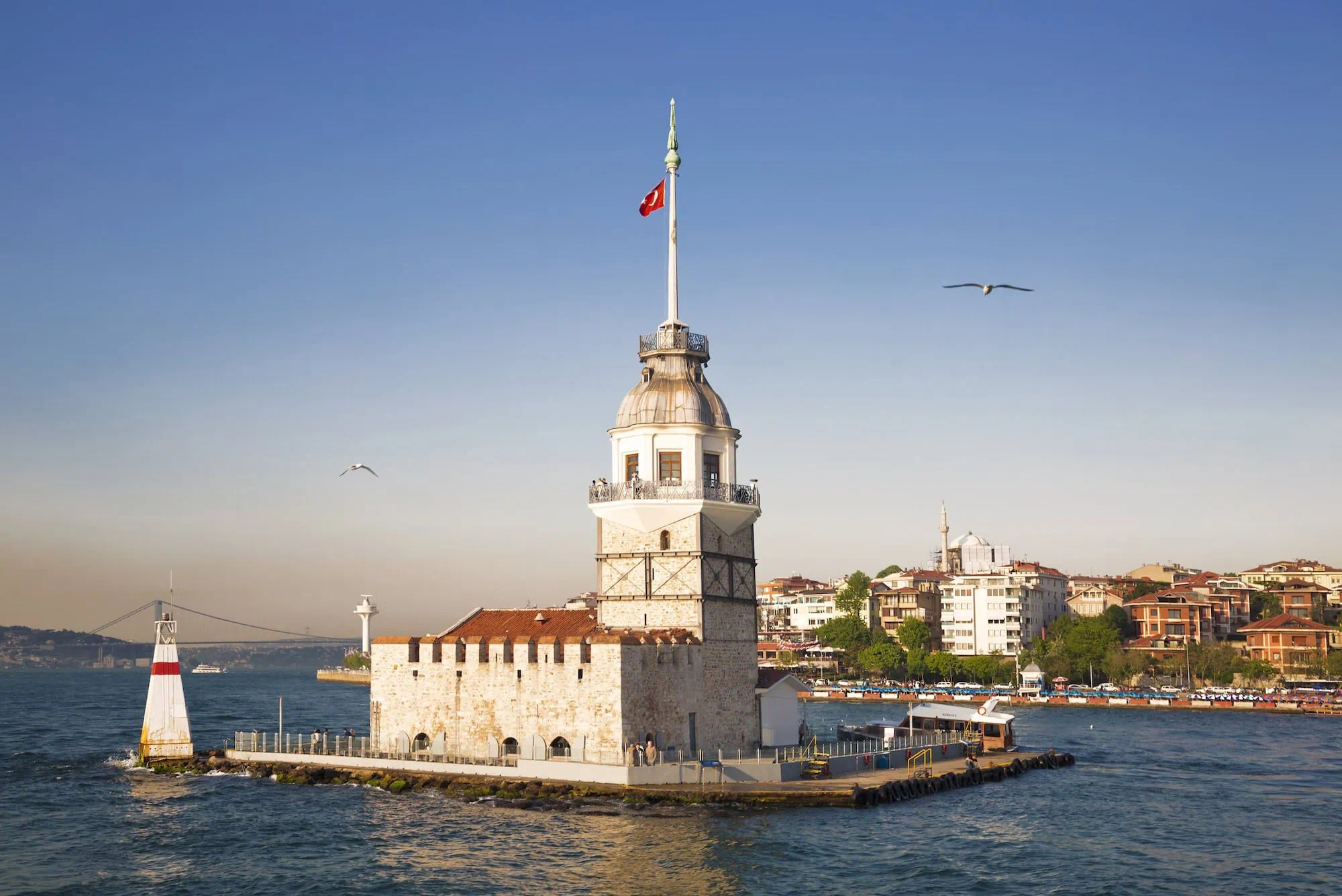Maiden's Tower illuminated in golden light with Turkish flag and seagulls flying overhead