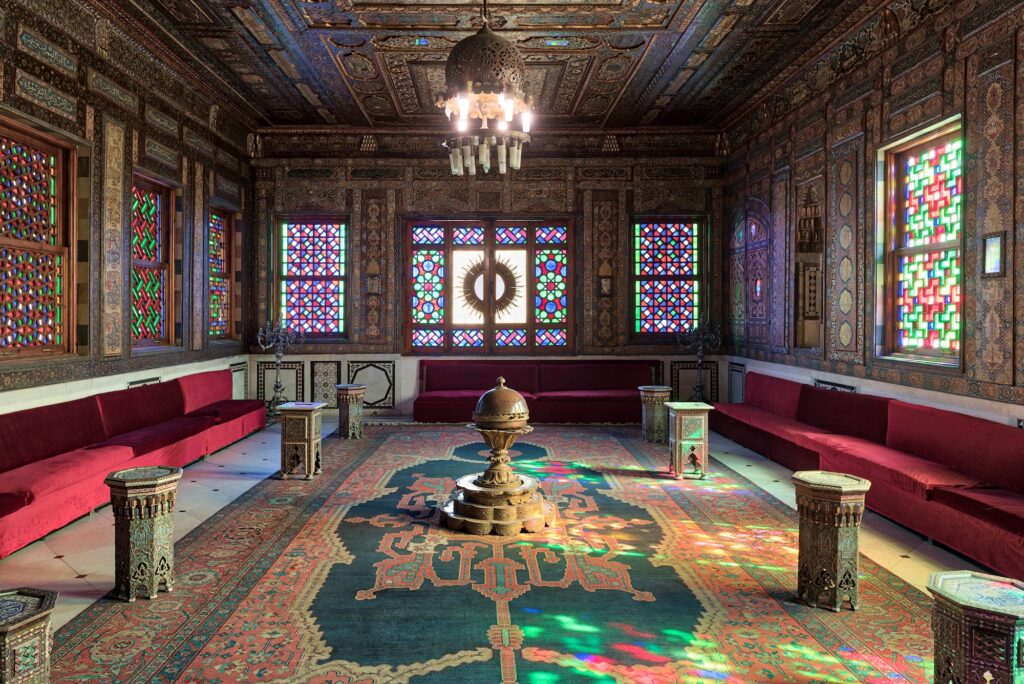 The Syrian Hall interior with carved wooden walls and ceiling, stained glass windows, Ottoman emblem, and hanging chandelier at Manial Palace, Cairo