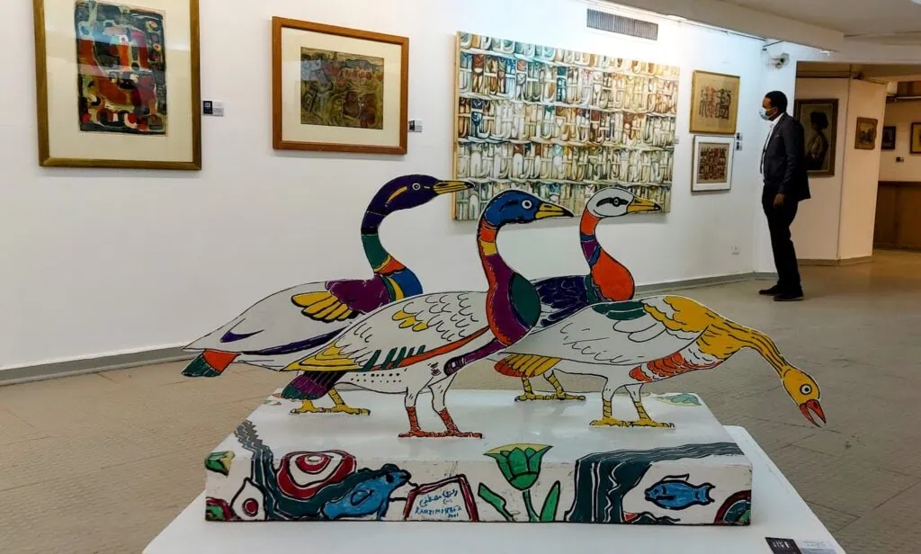 An interior gallery view with a colorful bird sculpture display, framed modern artworks on white walls, and a visitor viewing exhibits at the Museum of Modern Art, Cairo