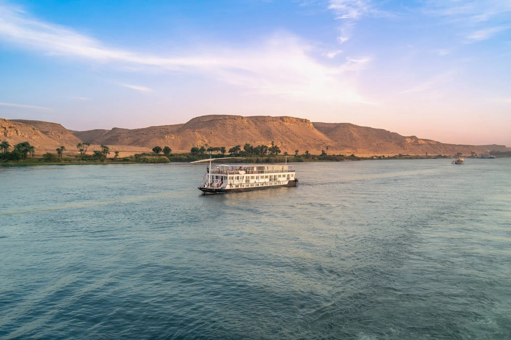 Modern Nile River cruise boat sailing past desert cliffs and palm trees