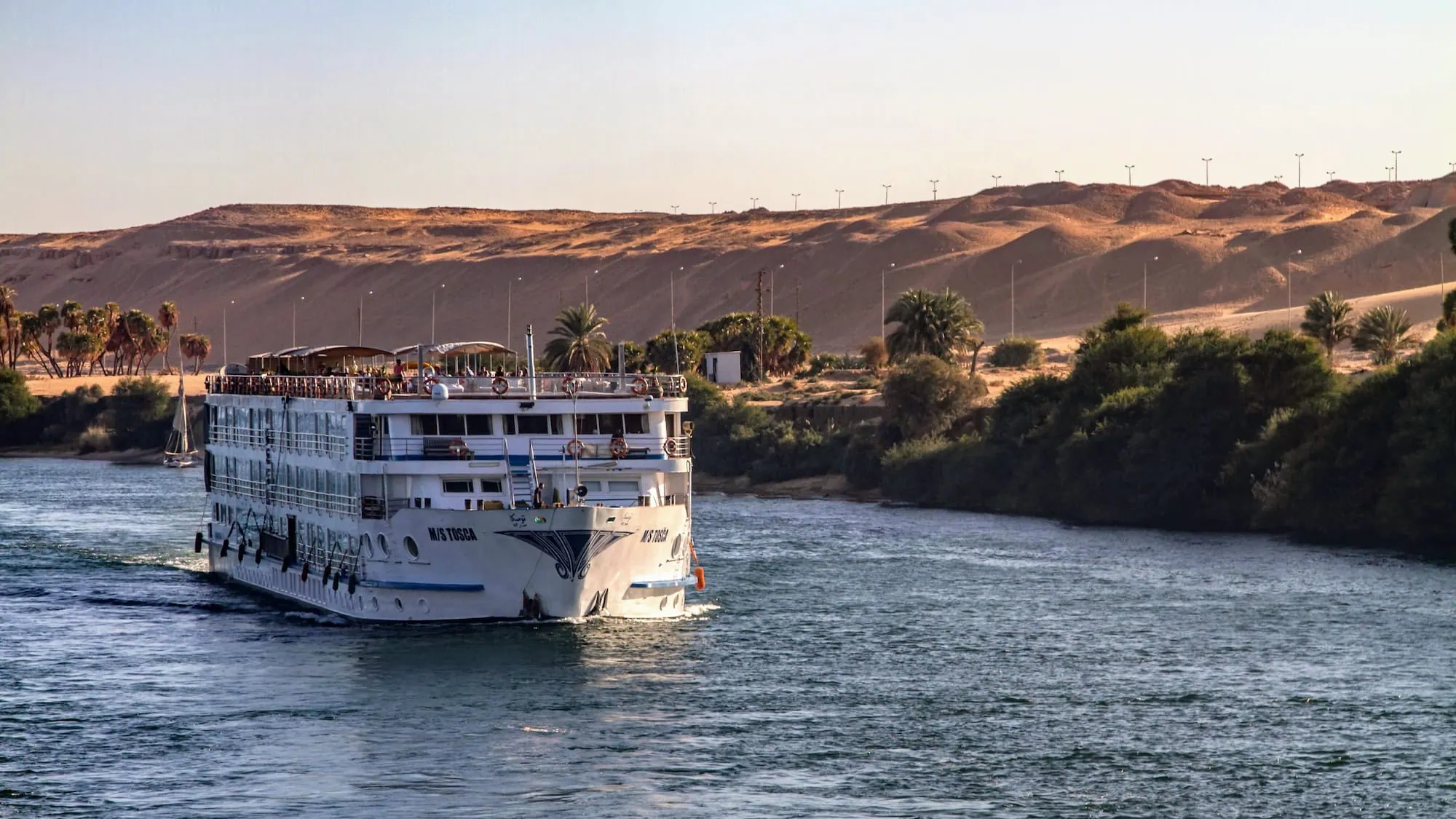 White multi-deck cruise ship sailing on the Nile River near Aswan with desert hills and palm trees