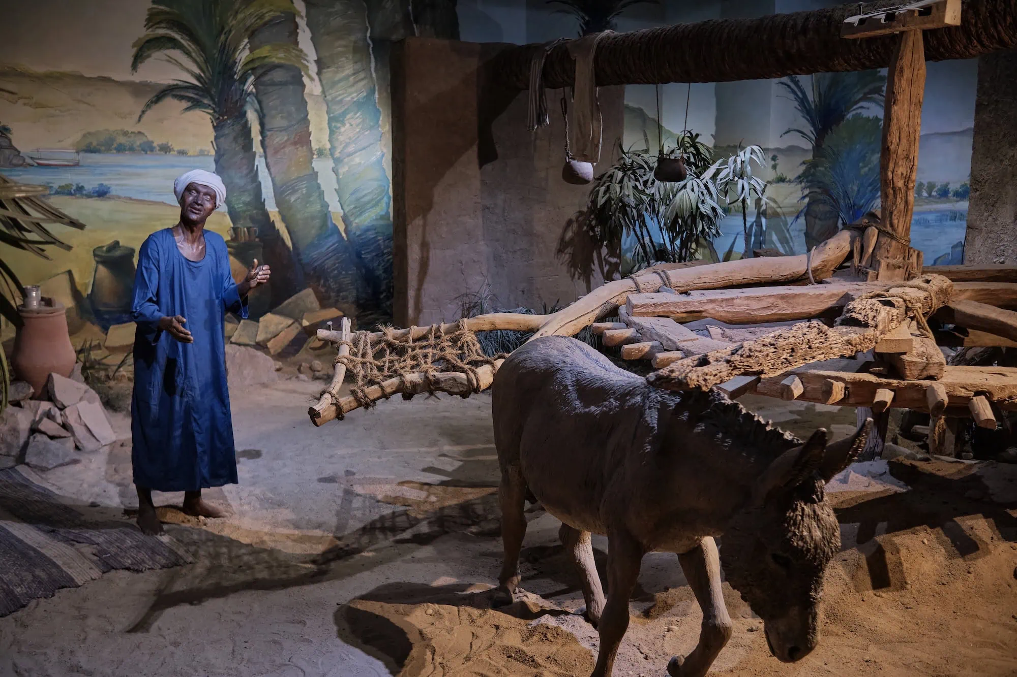 Exposição de museu mostrando aldeia tradicional egípcia com búfalo d'água e carrinho de madeira