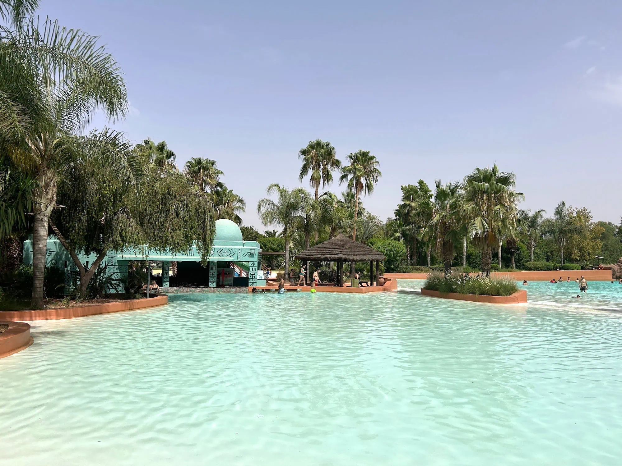 Tropical water park with large swimming pool surrounded by palm trees and thatched umbrellas