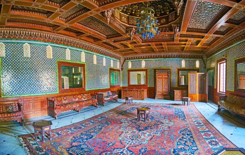A panoramic interior view with carved wooden ceiling, patterned wall tiles, and architectural details inside the Moroccan Hall of the Manial Palace, Cairo