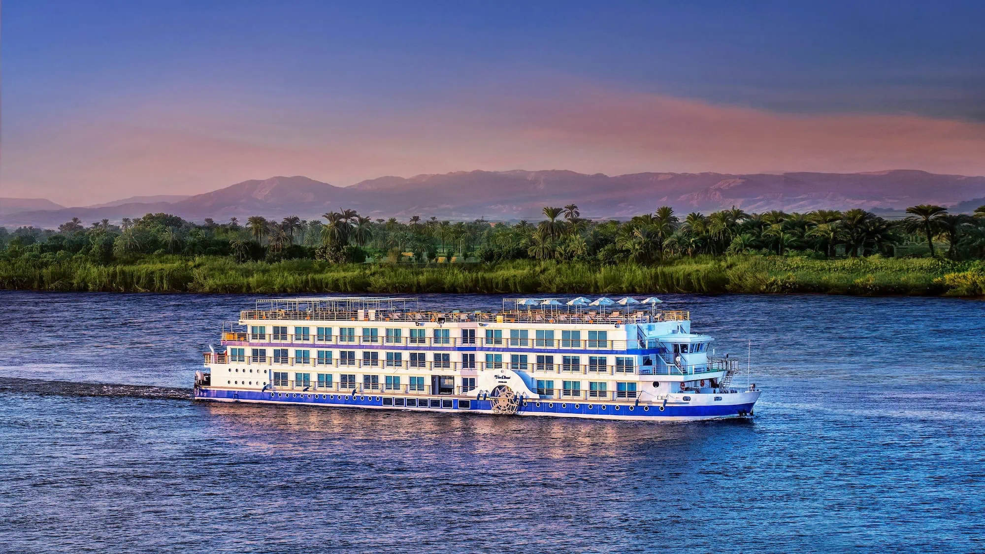 Nile cruise ship sailing on the river at sunset with palm trees along the shore