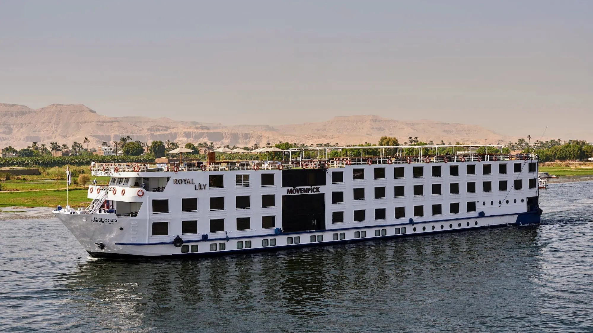 Luxury cruise ship sailing on the Nile River past desert mountains and palm trees