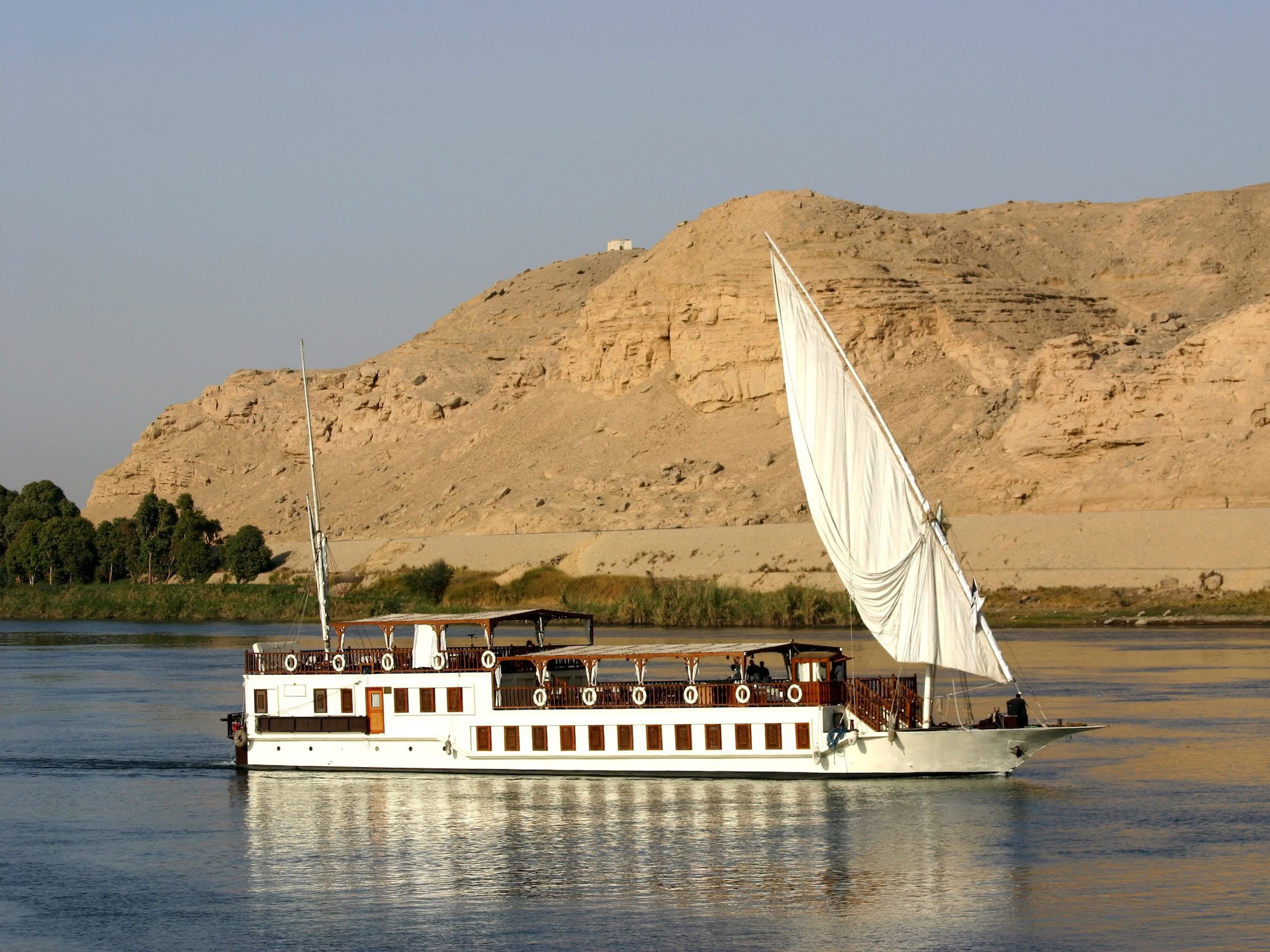 Traditional sailing boat with white sail on the Nile River surrounded by desert cliffs