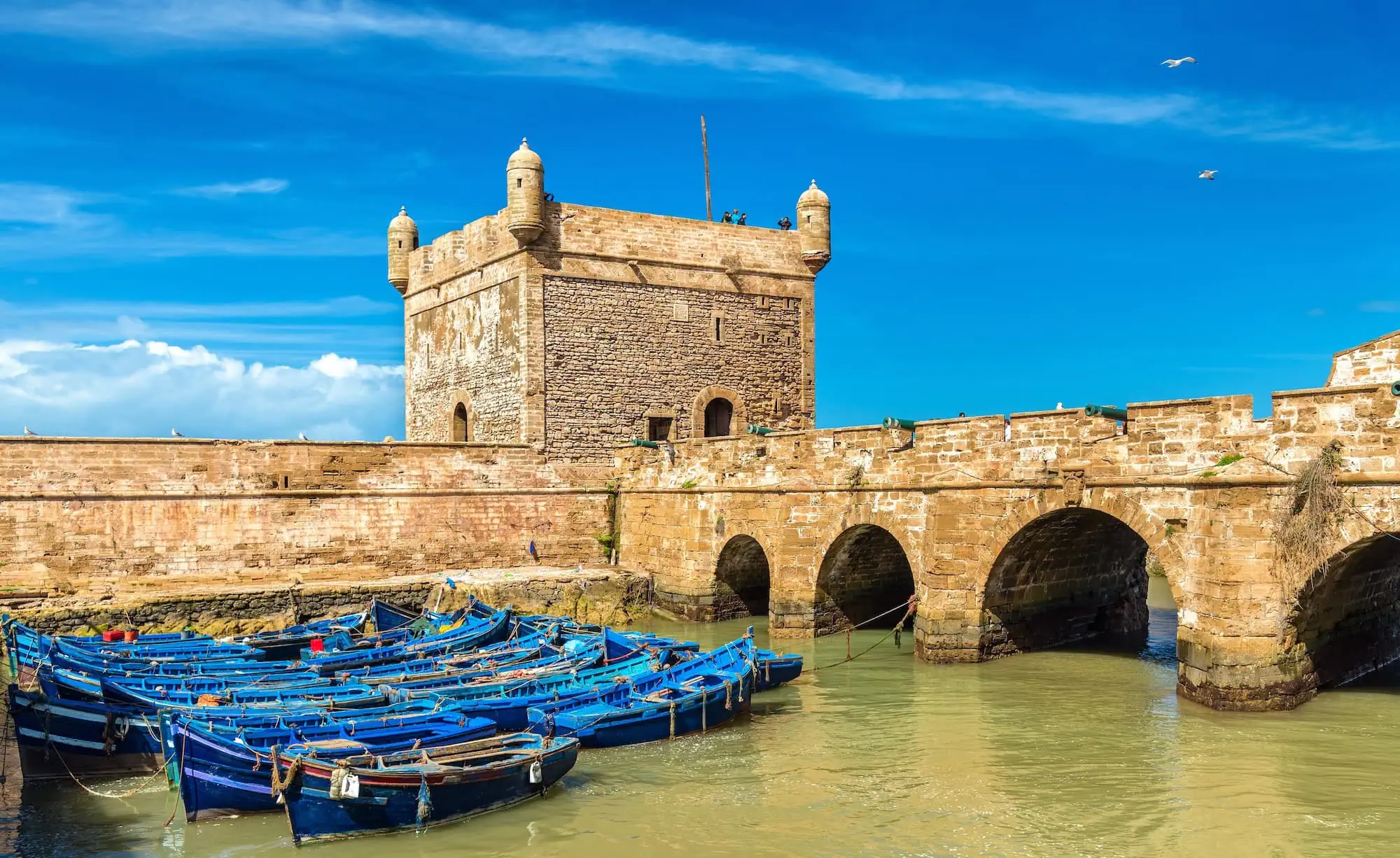 Historic Sqala du Port fortress tower and stone walls overlooking traditional blue fishing boats in Essaouira harbor