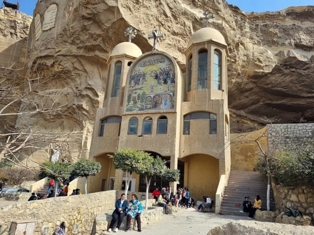 An exterior view showing tourists near the rock-cut facade and cave entrance of Saint Simon the Tanner Church, Cairo