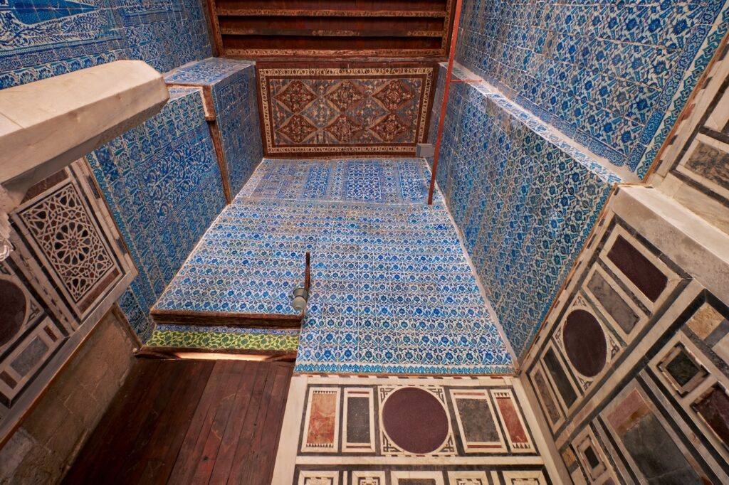 Decorated interior walls and upper loft with blue tilework, stone arches, and geometric patterns inside the Aqsunqur Mosque, Cairo