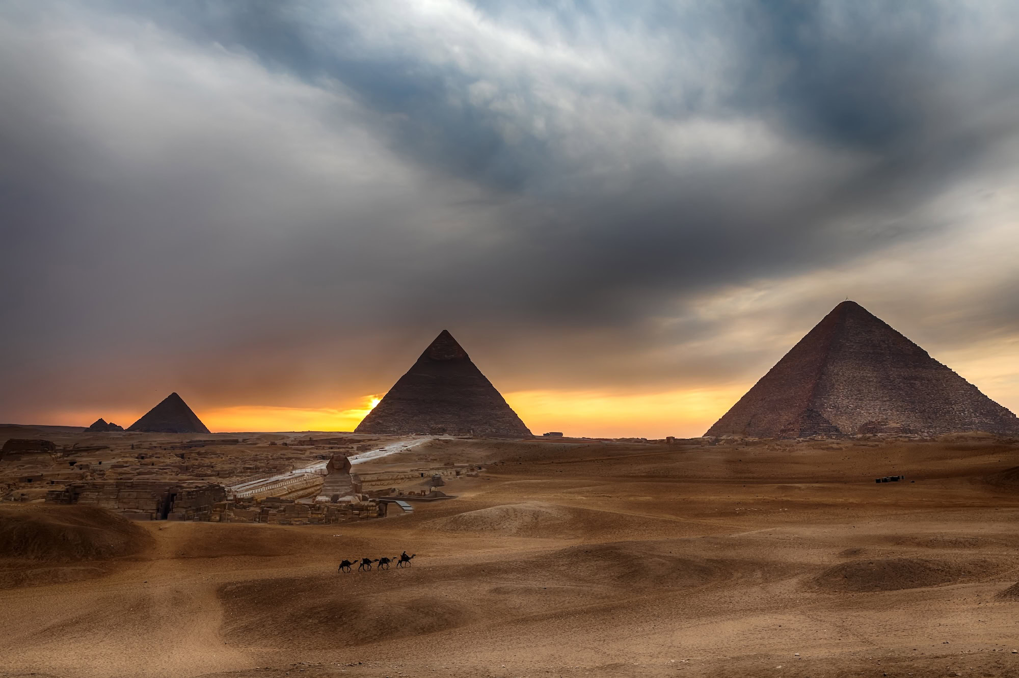 Great Pyramids of Giza and Sphinx at sunset with camel caravan in desert landscape