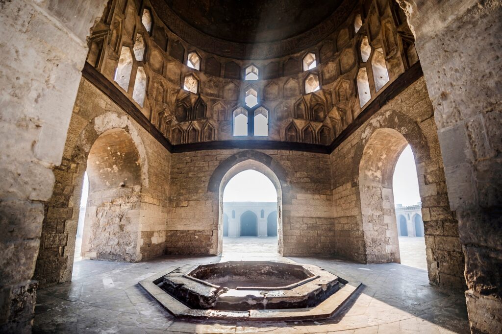 A domed interior pavilion with stone arches, carved window openings, and a central ablution fountain at the Ibn Tulun Mosque, Cairo