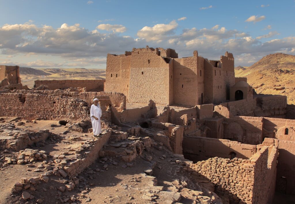 Ruins of St. Simeon Monastery with an Egyptian man in traditional robe standing in front under a partly cloudy sky, Aswan