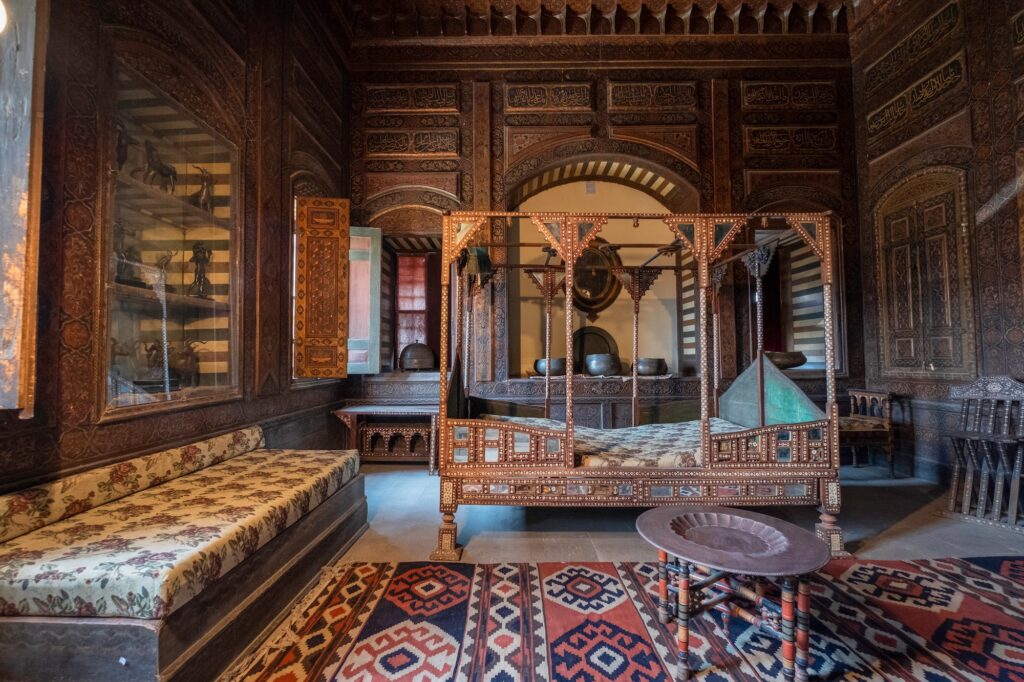 An interior room with carved wooden walls, inlaid wooden bed, traditional furnishings, and patterned rugs inside the Gayer-Anderson Museum, Cairo