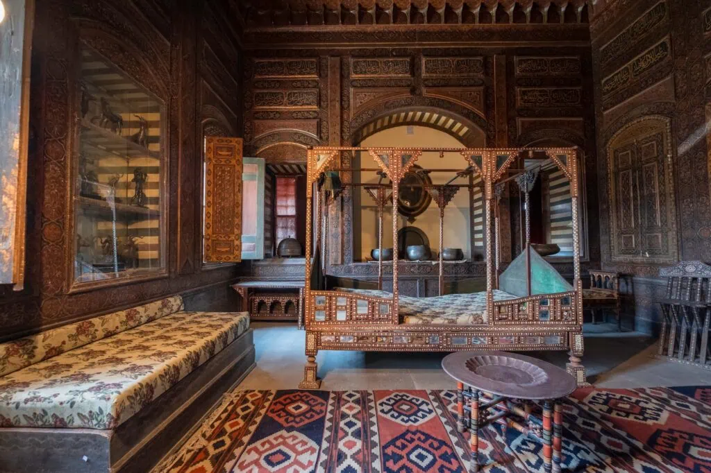 An interior room with carved wooden walls, inlaid wooden bed, traditional furnishings, and patterned rugs inside the Gayer-Anderson Museum, Cairo