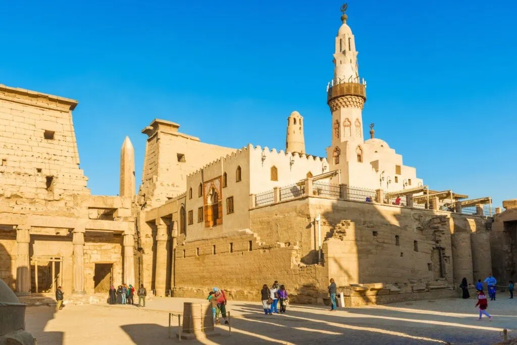 Visitors walking among stone columns with carved reliefs and the Abu Haggag Mosque within the Luxor Temple complex, Luxor