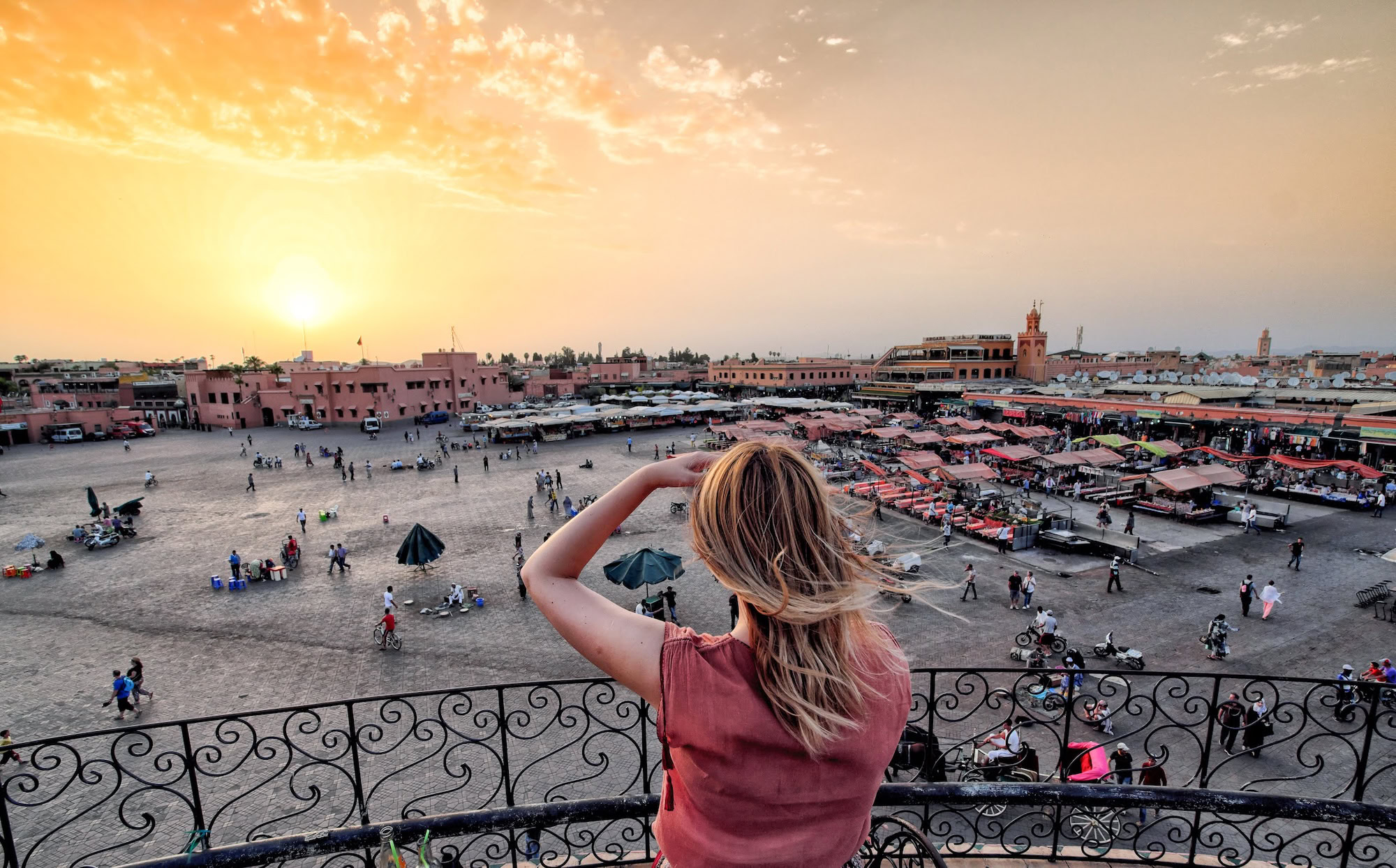 Woman on balcony overlooking bustling Jemaa el-Fnaa square in Marrakech with mosque minaret