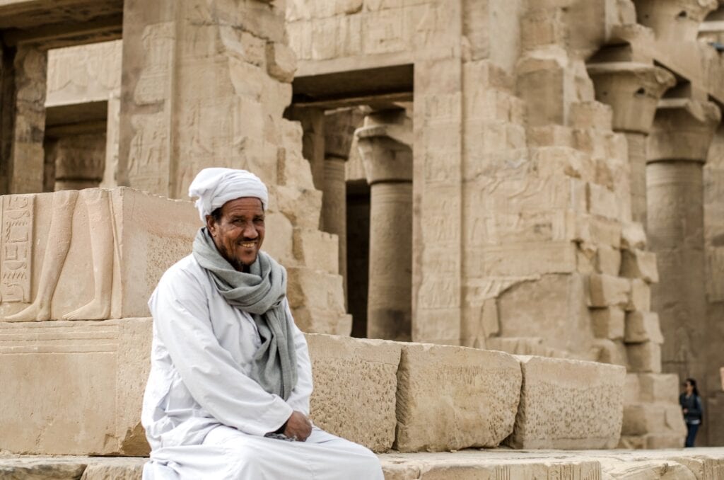 Traditional temple guard sitting in front of an ancient pharaoh temple with stone columns and carvings, Aswan
