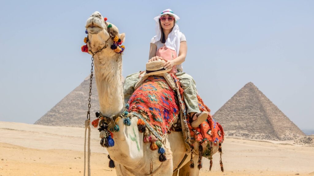 Woman riding a camel across the desert landscape with pyramids visible in the background, Giza