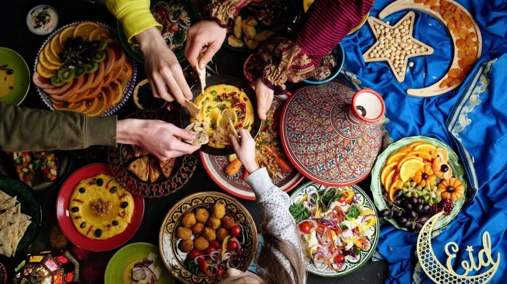 Overhead view of hands sharing an iftar table of hummus, falafel, flatbread, and fruit platters during Ramadan, with an Eid crescent decoration beside the dishes.